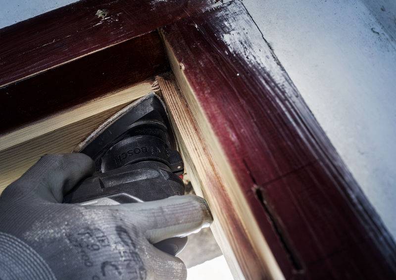 Een hand met een handschoen houdt een elektrisch gereedschap vast en zaagt hout aan een rood gelakt raamkozijn.