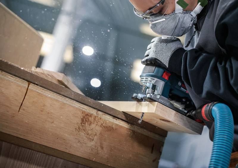 Een persoon met veiligheidsbril en mondmasker gebruikt een elektrisch gereedschap om een houten plank te zagen in een werkplaats.