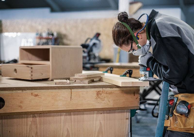 Een persoon draagt veiligheidsbril en gehoorbescherming terwijl hij hout bewerkt aan een werktafel in een timmerwerkplaats.
