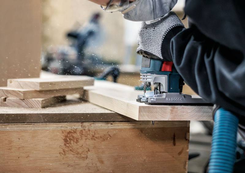 Een persoon werkt met een bovenfrees aan een houten plank; houtspaanders vliegen door de lucht. Beschermende handschoenen en gereedschap zijn zichtbaar.