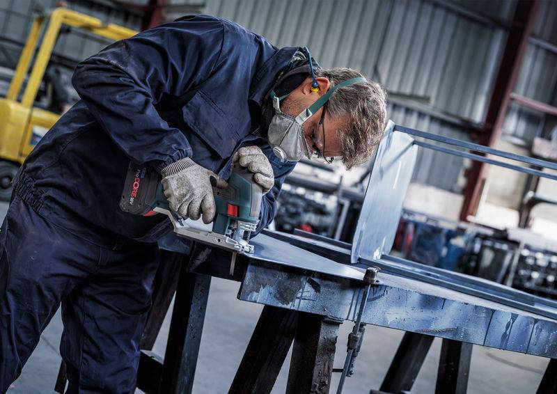 Een persoon zaagt een metalen stuk met een elektrische zaag in een werkplaats. Ze draagt een beschermingspak en een adembeschermingsmasker.