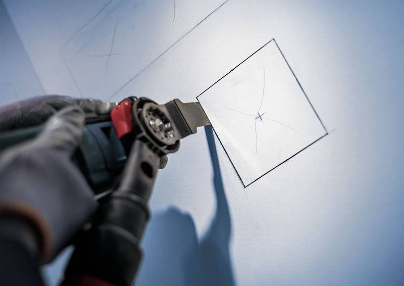 Een persoon snijdt nauwkeurig een vierkant gat in een muur met een elektrisch gereedschap, vermoedelijk voor een installatie.