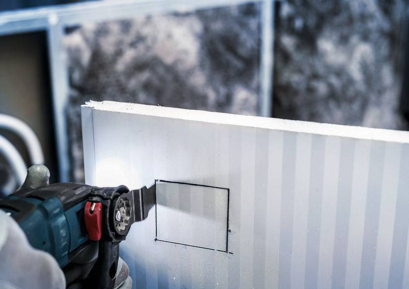 Een persoon zaagt met een kettingzaag een rechthoekig gat in een metalen wand, omgeven door een bouwplaatsomgeving.