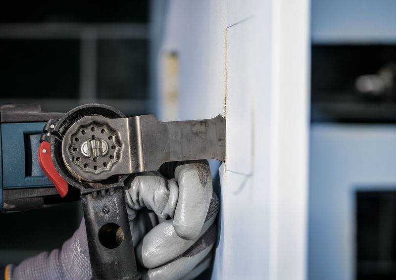 Een hand met werkhandschoenen bedient een oscillerend gereedschap dat een rechthoekige vorm uit het pleisterwerk van de muur snijdt.