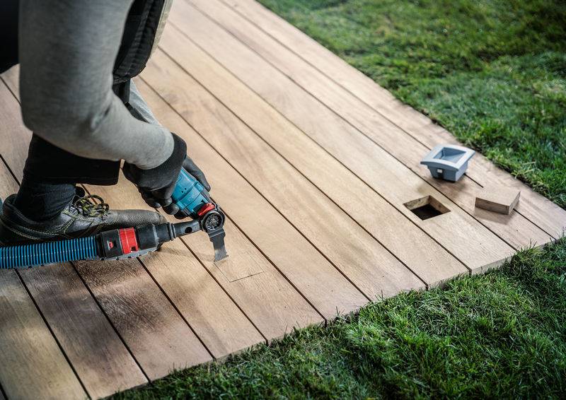 Een persoon snijdt een rechthoekig gat in een houten plank op een gazon. Naast het gat ligt een open elektrische doos.