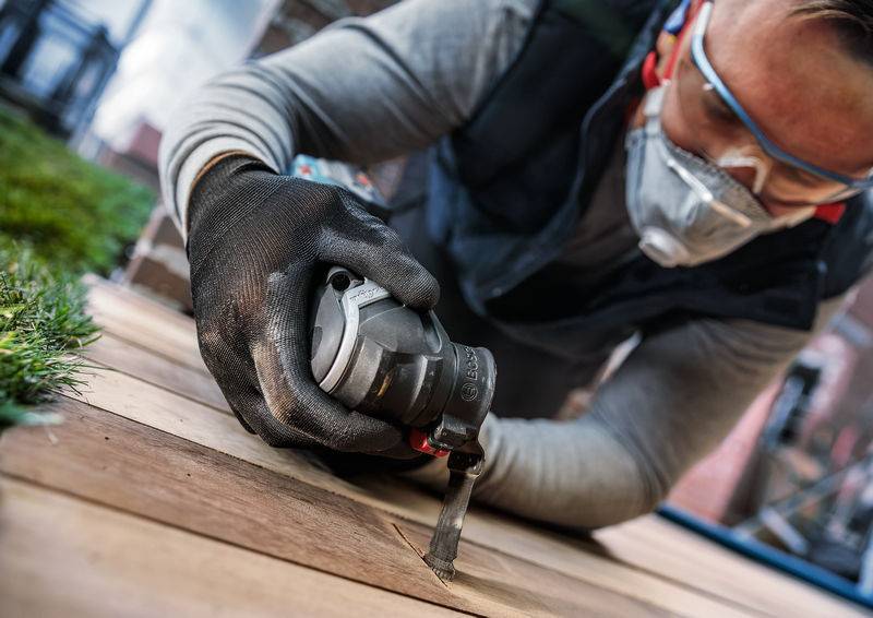 Persoon die veiligheidsbril en masker draagt, gebruikt een elektrisch gereedschap om houten planken buiten te bewerken.