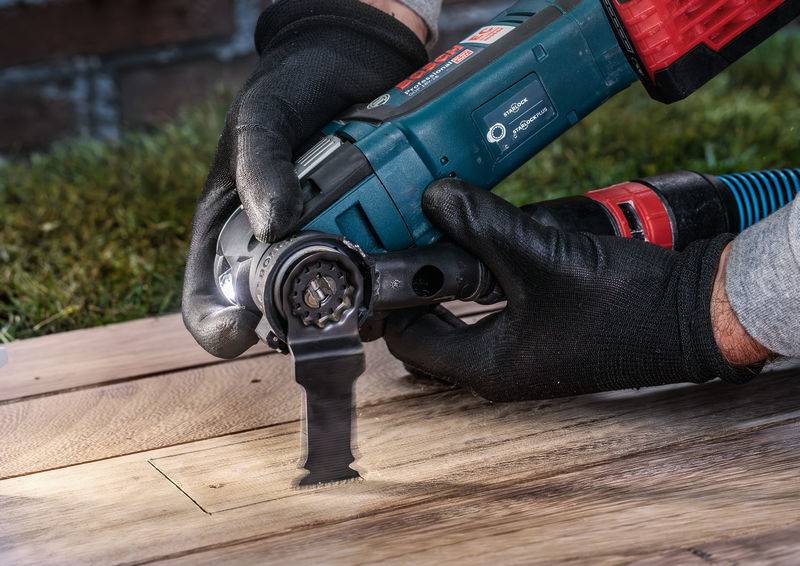 Een persoon werkt met een oscillerend multitool op een houten vloer. Het gereedschap heeft zwarte zaagbladen. Handen in zwarte handschoenen.