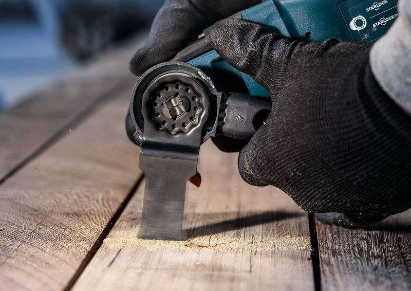 Een ambachtsman draagt zwarte handschoenen en gebruikt een elektrisch gereedschap om hout te zagen. Fijn houtstof wordt opgewaaid.