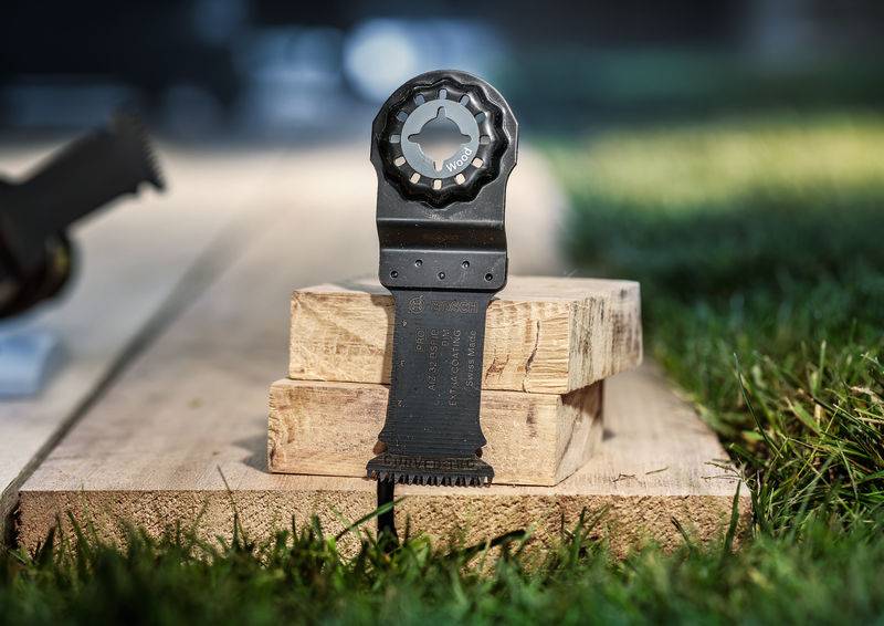 Een multitool-mes staat op gestapelde houten planken buiten. Het is ontworpen voor precisiewerk in hout en nagels.