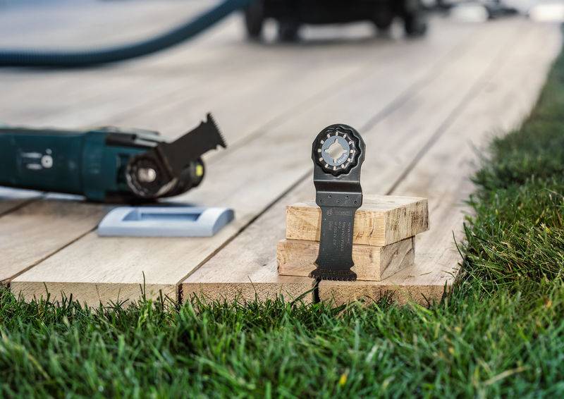 Een elektrische zaag met een houtsnijblad ligt op een houten terras, naast gezaagde houten stukken en gras.