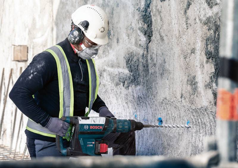 Arbeider met helm en veiligheidsbril boort met een Bosch-boormachine in een betonnen muur. Veiligheidsuitrusting en werkkleding zijn aanwezig.