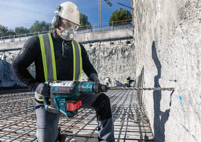 Arbeider met veiligheidshelm en masker gebruikt een boorhamer om een groot betonnen fundament te bewerken; bouwplaats op de achtergrond.