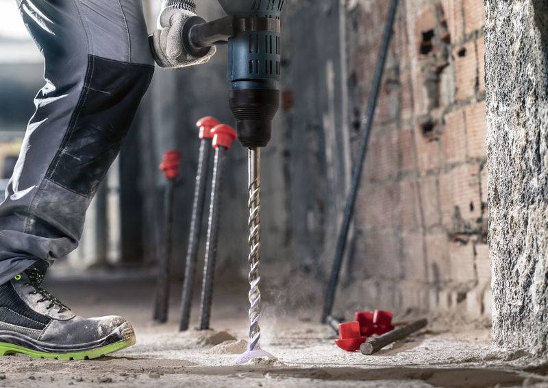 Een persoon boort met een elektrische boormachine in een betonnen vloer. Op de achtergrond zijn metalen staven en rode markeringen aan de muur zichtbaar.