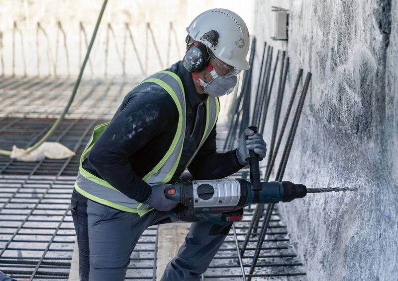 Werknemer in beschermende uitrusting boort in betonnen muur op bouwplaats. Gereedschap in actie, omgeving toont stalen balken en betonnen vloer.