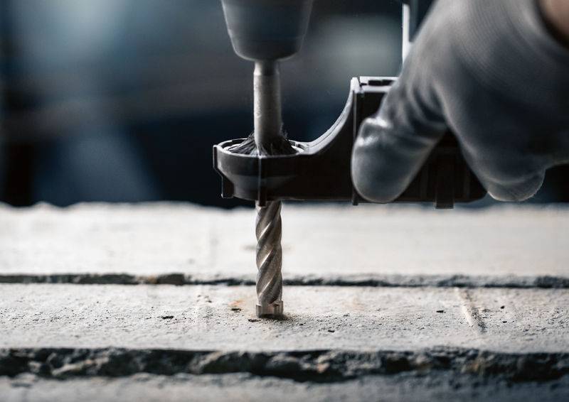 Hand met handschoen houdt een boormachine vast terwijl hij een gat boort in een betonoppervlak. Stofdeeltjes zijn zichtbaar.