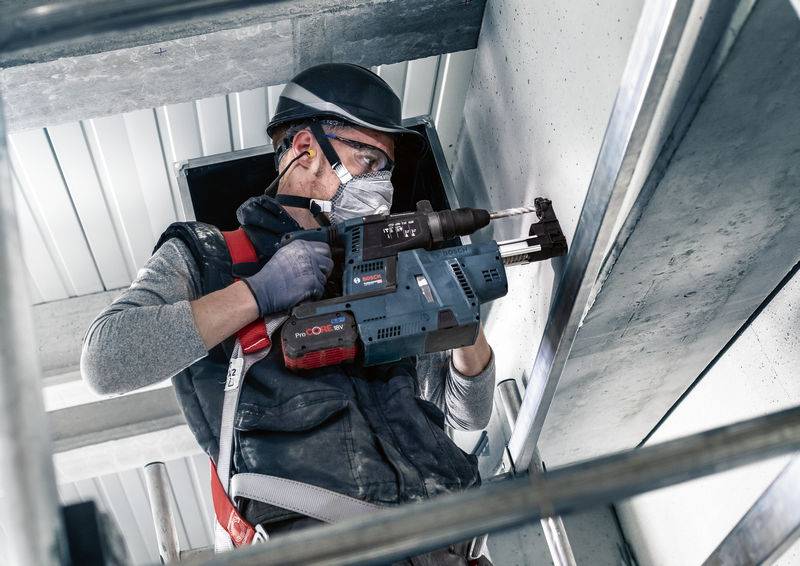 Een persoon in beschermende kleding gebruikt een boorhamer om te werken in een industriële omgeving. Veiligheidsbril en helm worden gedragen.