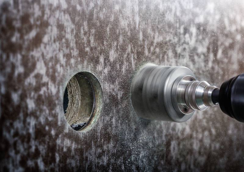 Close-upfoto van een boor die een groot gat boort in een metalen plaat. Links is een reeds geboord gat zichtbaar.