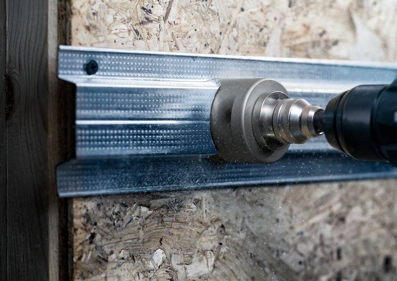 Een close-up toont een boormachine die een gat boort in een metalen profiel op een spaanplaat, omlijst door gestructureerd hout.