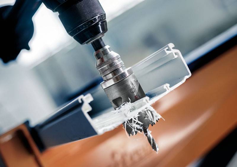 Een metalen boor doorboort een stuk wit plastic, spaanders vliegen weg. Opgenomen in een industriële omgeving.