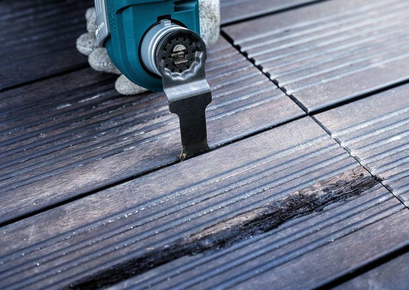 Een handmatige machine snijdt een nauwkeurige sleuf in een houten terrasplank. Een persoon draagt handschoenen tijdens het werk.