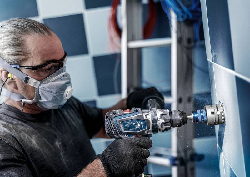 Een man met veiligheidsbril en masker boort tegels in een badkamer. Op de achtergrond is een ladder zichtbaar.