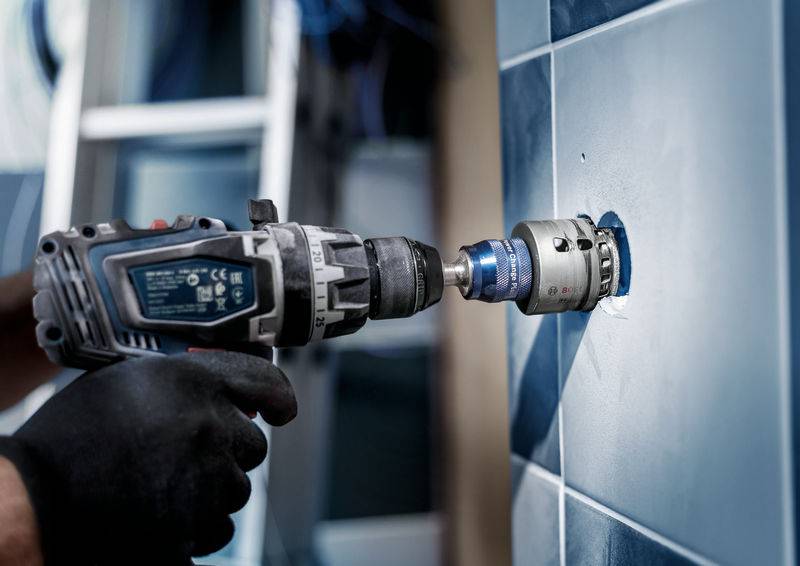 Een persoon boort een gat in een blauw betegelde muur met een boormachine. Focus op de boor en de tegels.