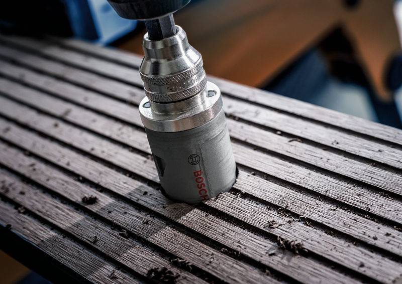Een metalen boor met Bosch-logo boort in een geprofileerd houten bord. Houtkrullen zijn verspreid rond de boorlocatie.
