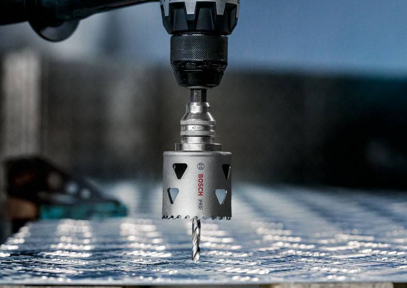 Een boormachine met een gatzaagopzetstuk boort een rond gat in een metalen plaat. Op de achtergrond wazig gereedschap.