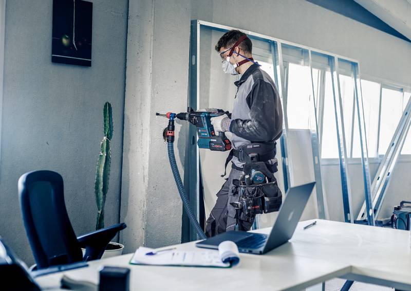 Een ambachtsman werkt met een boormachine in een kantoor. Hij draagt beschermende kleding en een adembeschermingsmasker. Op tafel ligt een laptop.