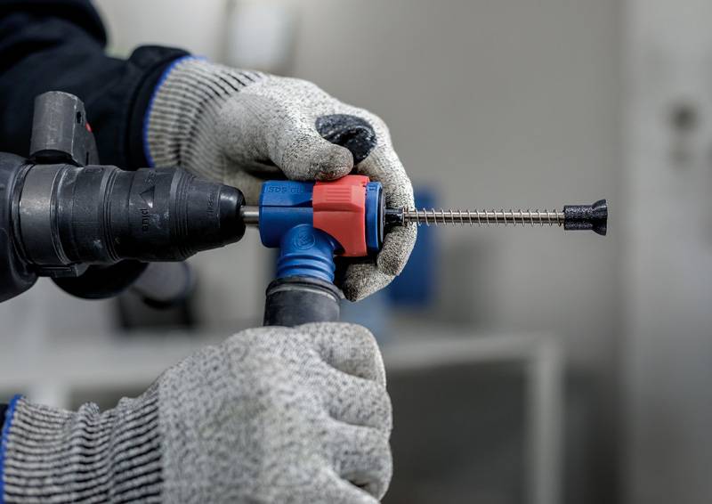 Een persoon draagt grijze handschoenen en houdt een boormachine vast met een blauwe en rode boorbit.