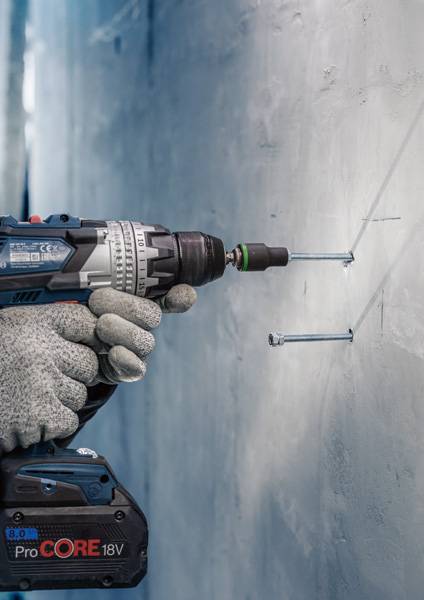 Een persoon boort met een accuschroefmachine in een betonmuur en bevestigt schroeven. De boormachine draagt een logo 'ProCORE 18V'.