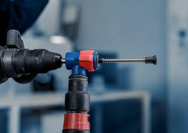 Een boormachine wordt gebruikt om een lange schroef te draaien in een blauwzwarte voorziening met een rode handgreep. Achtergrond onscherp.