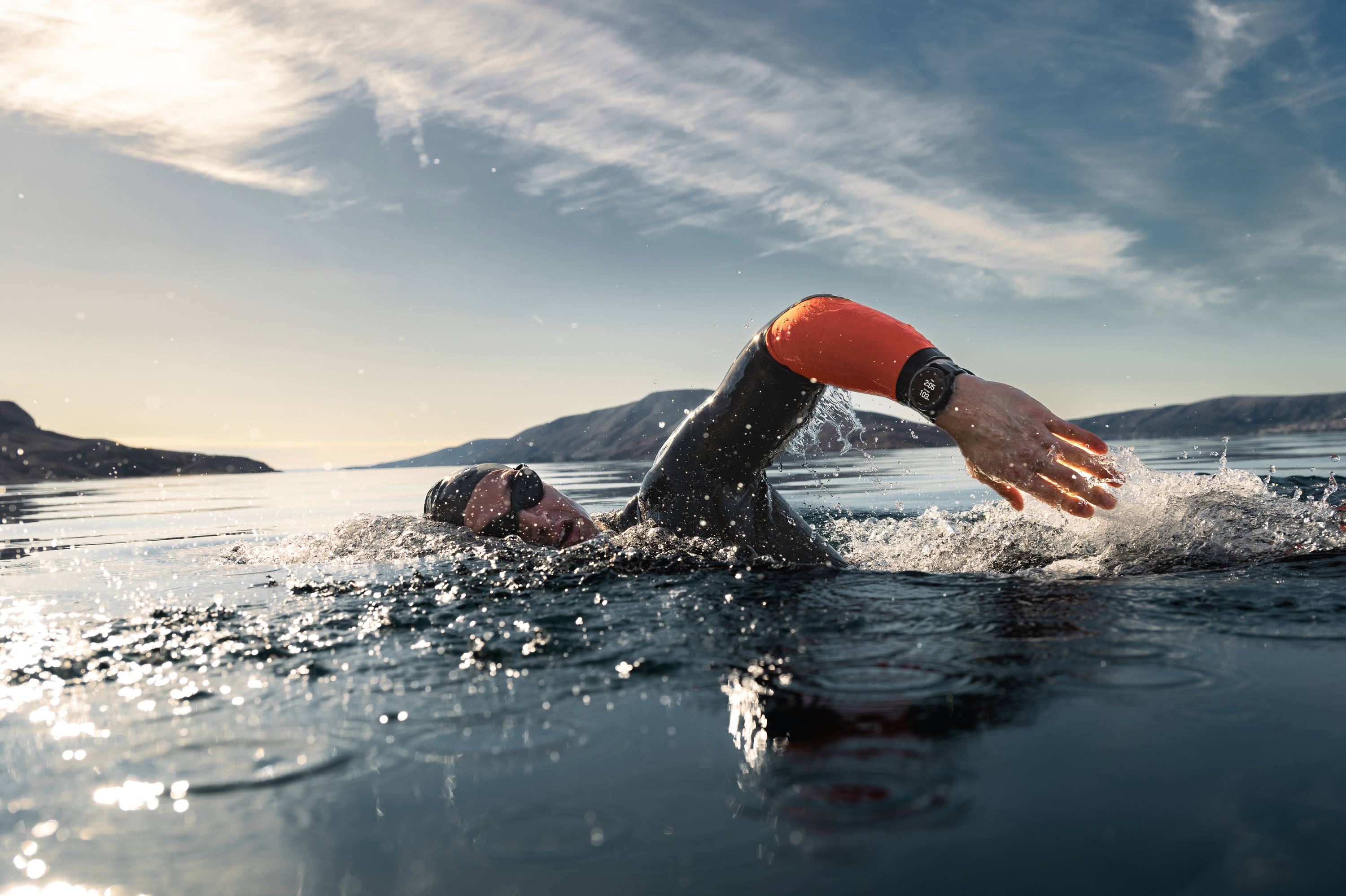 Een persoon zwemt in open water, draagt een neopreen wetsuit en een zwembril. Op de achtergrond is een bergachtig kustlandschap te zien.
