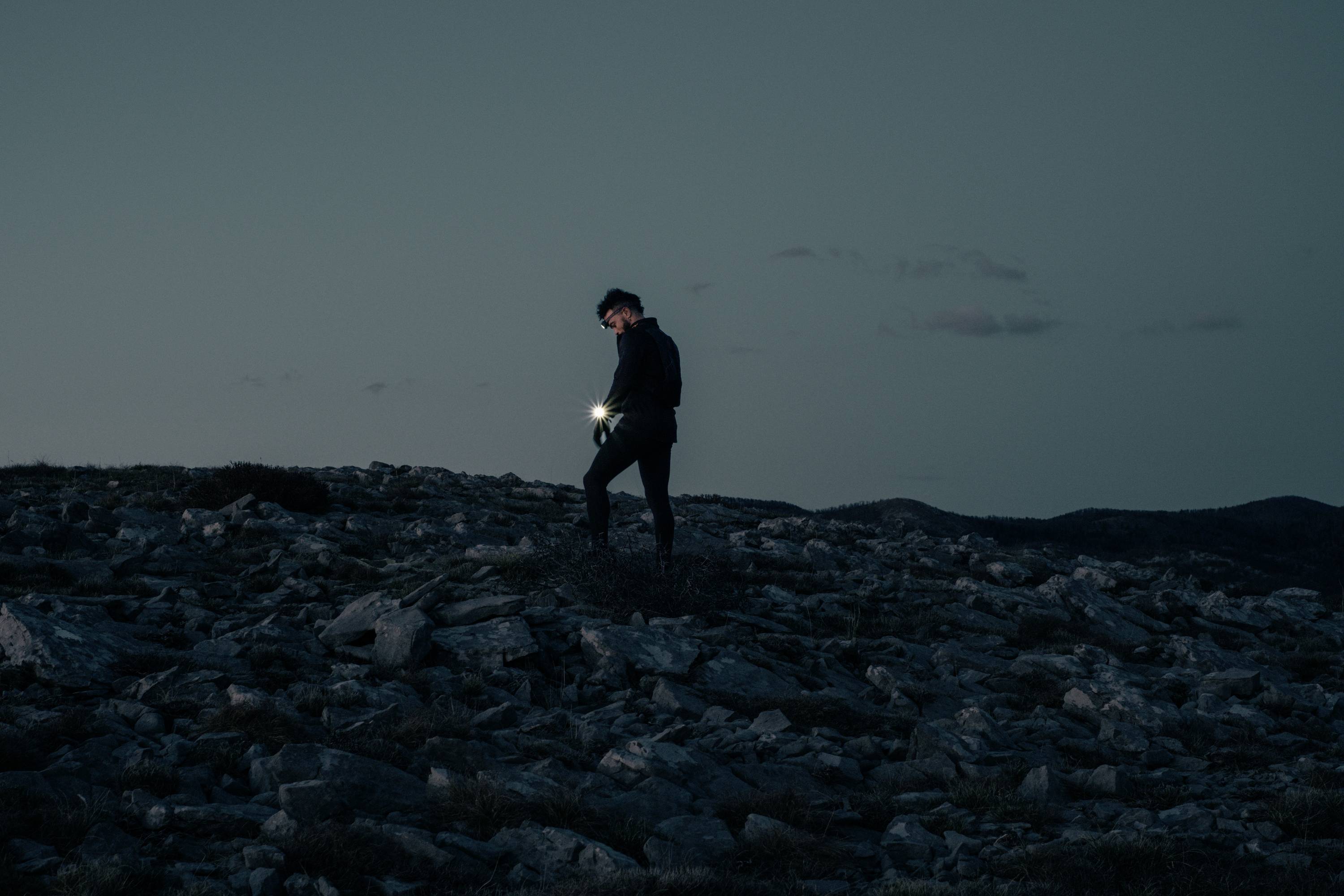Een persoon loopt bij schemering door een rotsachtig landschap, met een zaklamp in de hand die een deel van het pad verlicht.