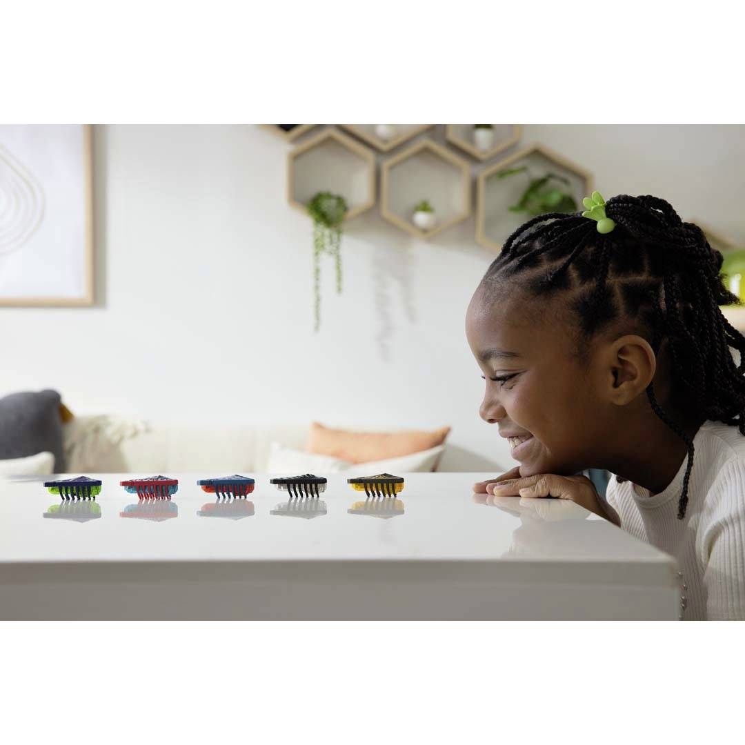 Een meisje met vlechten speelt met kleurrijke kevers op een tafel. De kevers staan op een rij. Op de achtergrond zijn plantenrekken.