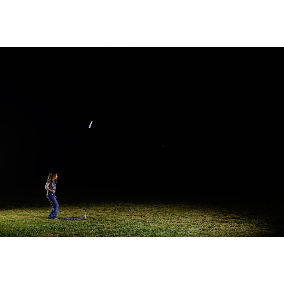 Een persoon kijkt 's nachts naar het lanceren van een modelraket op een veld. De raket licht op aan de hemel.