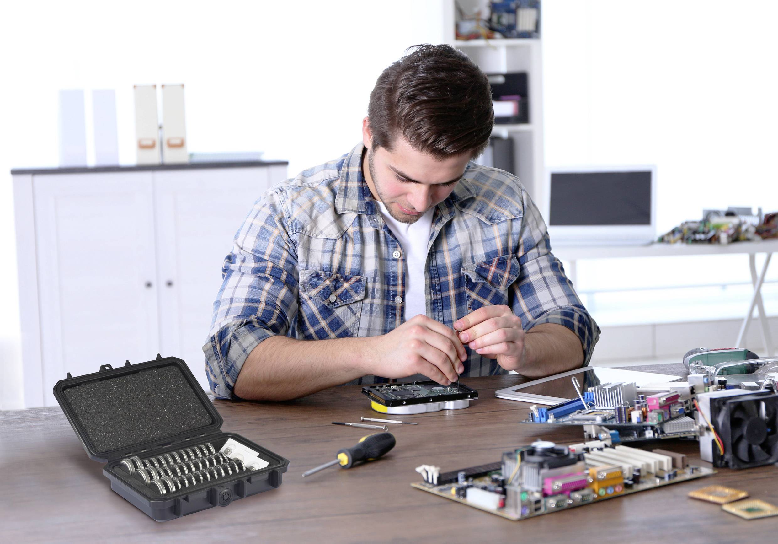Een man repareert een elektronisch apparaat aan een bureau. Op tafel liggen gereedschappen en elektronische componenten. Op de achtergrond zijn planken en toebehoren zichtbaar.