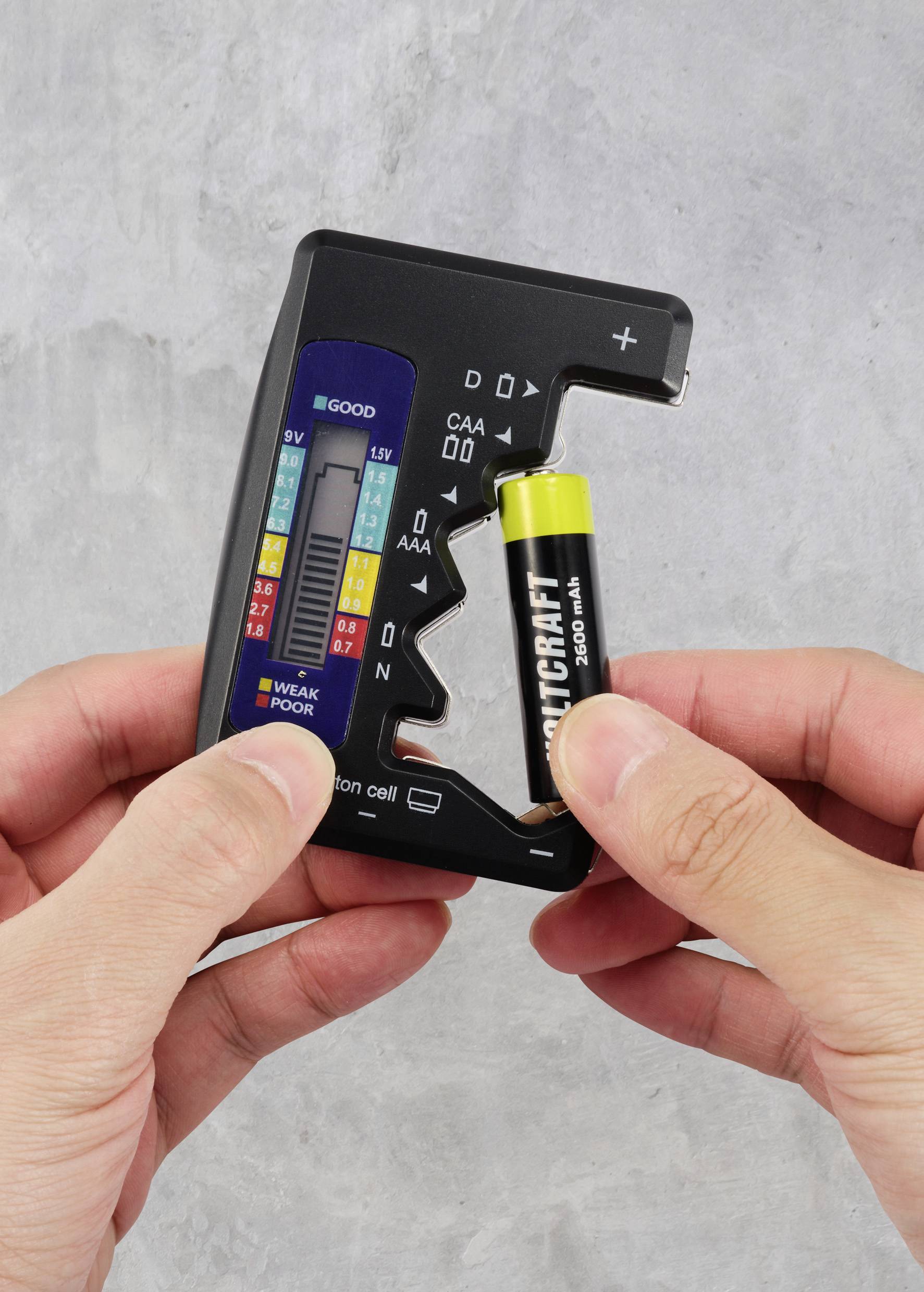 Een persoon houdt een batterijentester vast en test een AA-batterij. De tester geeft een goede prestatie aan. Grijze achtergrond.