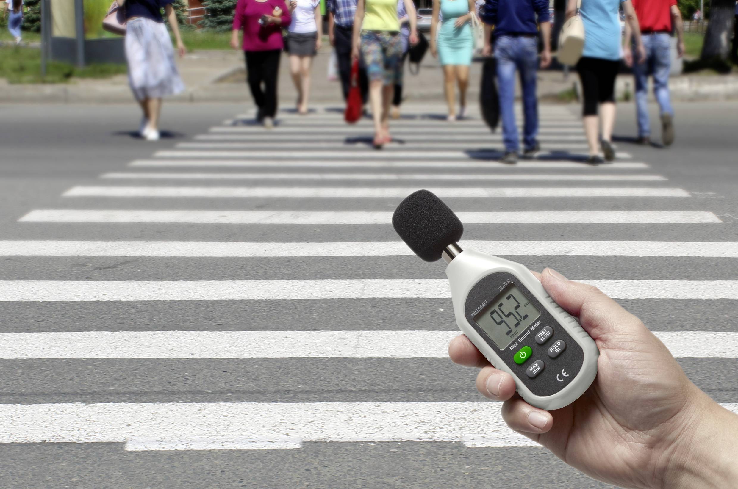 Een hand houdt een geluidsniveaumeter vast die 95,2 decibel aangeeft. Op de achtergrond steken mensen een zebrapad over.