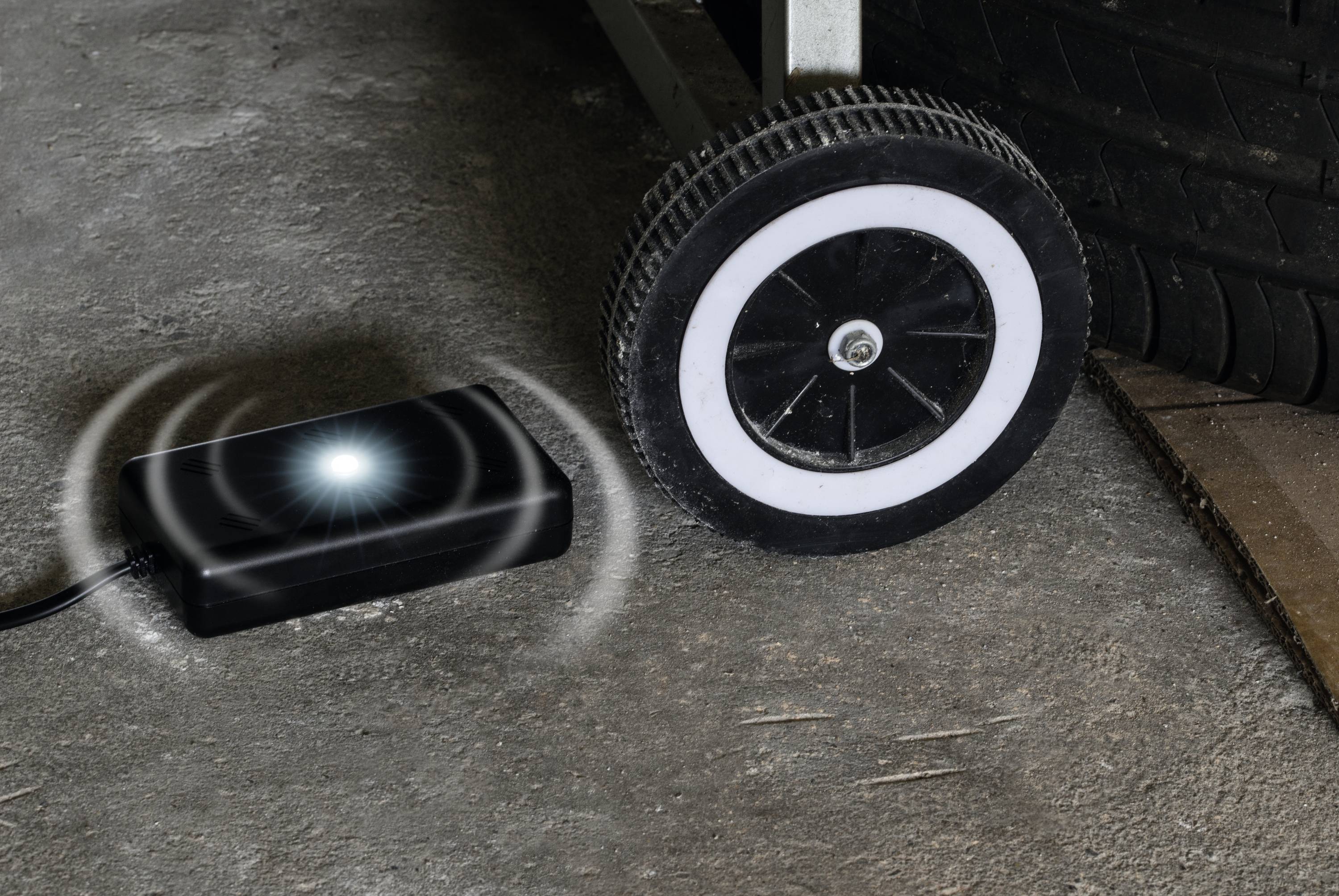 Een klein zwart apparaat met een oplichtende LED ligt op de grond naast het wiel van een voertuig, mogelijk bedoeld voor bewaking.