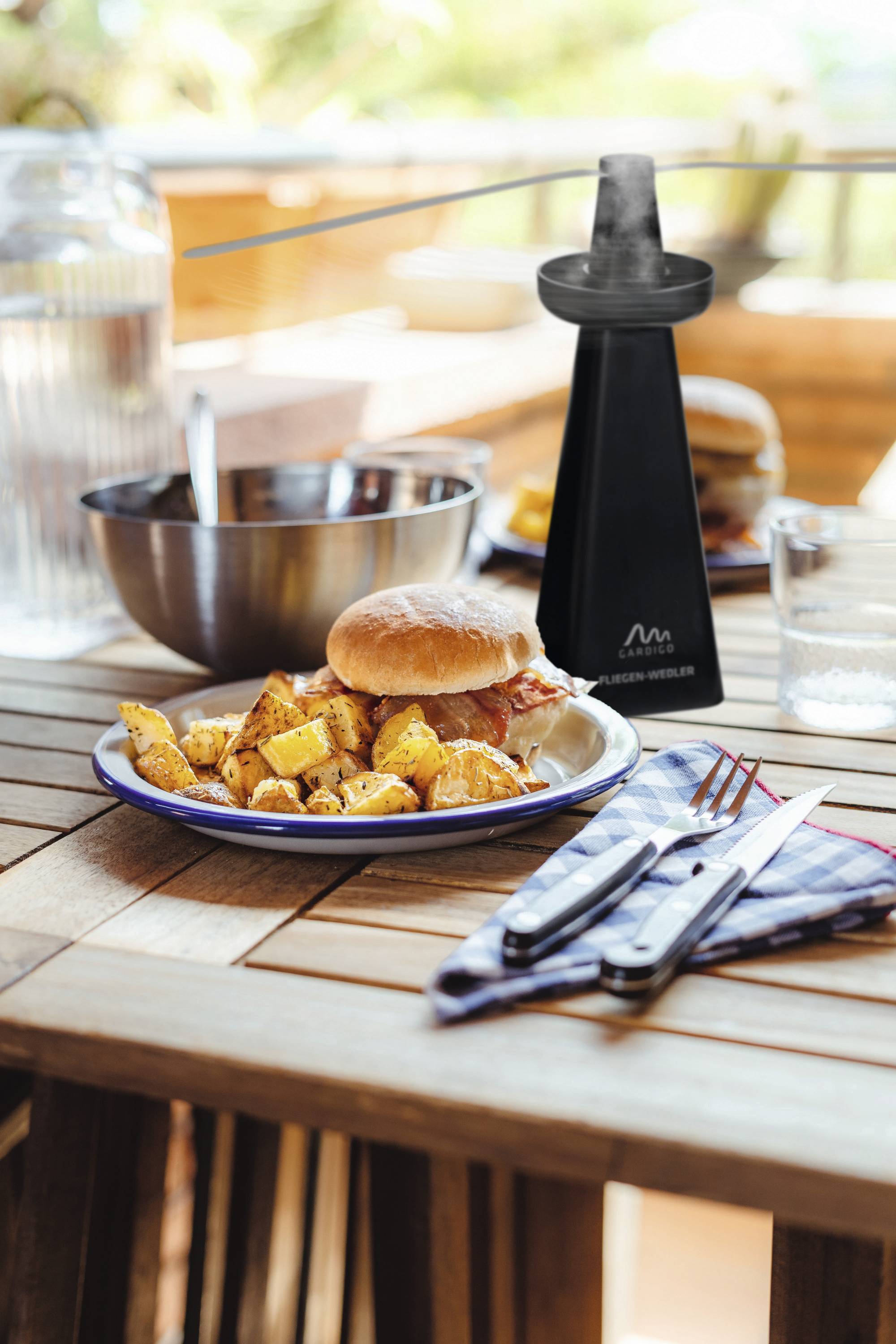 Een tafel op een terras met een bord friet en een hamburger, ernaast een waterkan, een metalen kom en een glas.