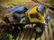 Twee op afstand bestuurde terreinvoertuigen op een miniatuur houten pad; de gele vrachtwagen vervoert een kleinere blauwe truck op zijn laadruimte.