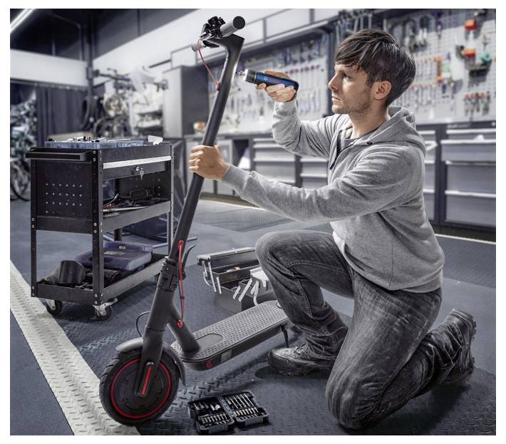 Een persoon repareert een elektrische step in een werkplaats. Ze houdt een gereedschap vast en werkt geconcentreerd aan het stuur.