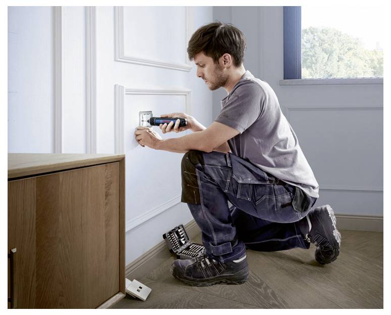 Een man repareert een stopcontact aan een muur. Hij knielt op de vloer, houdt een gereedschap in zijn hand, naast hem liggen andere gereedschappen.