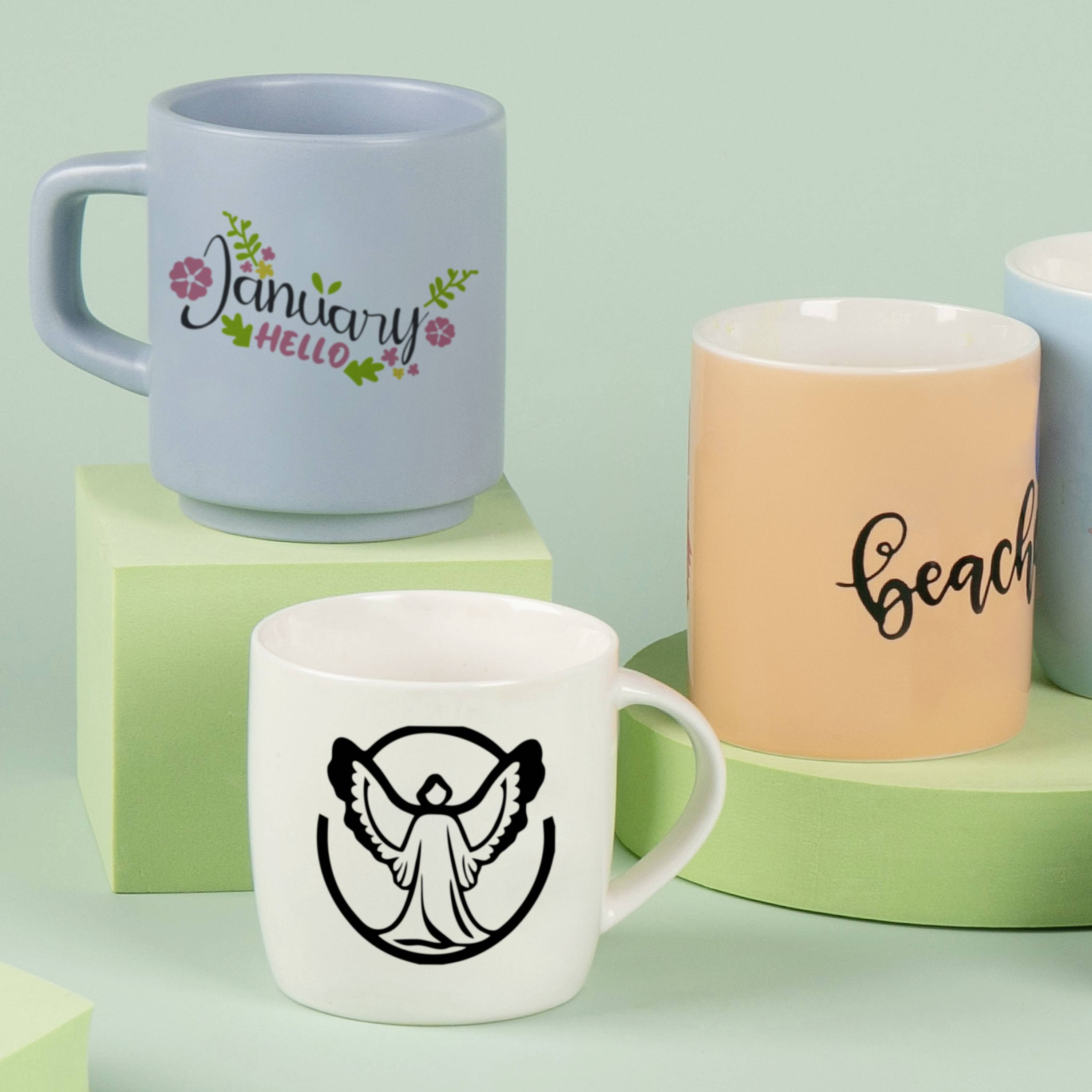 Drie kopjes op groene blokken: een blauwe met 'January Hello', een beige met 'Beach' en een witte met een engelenmotief.