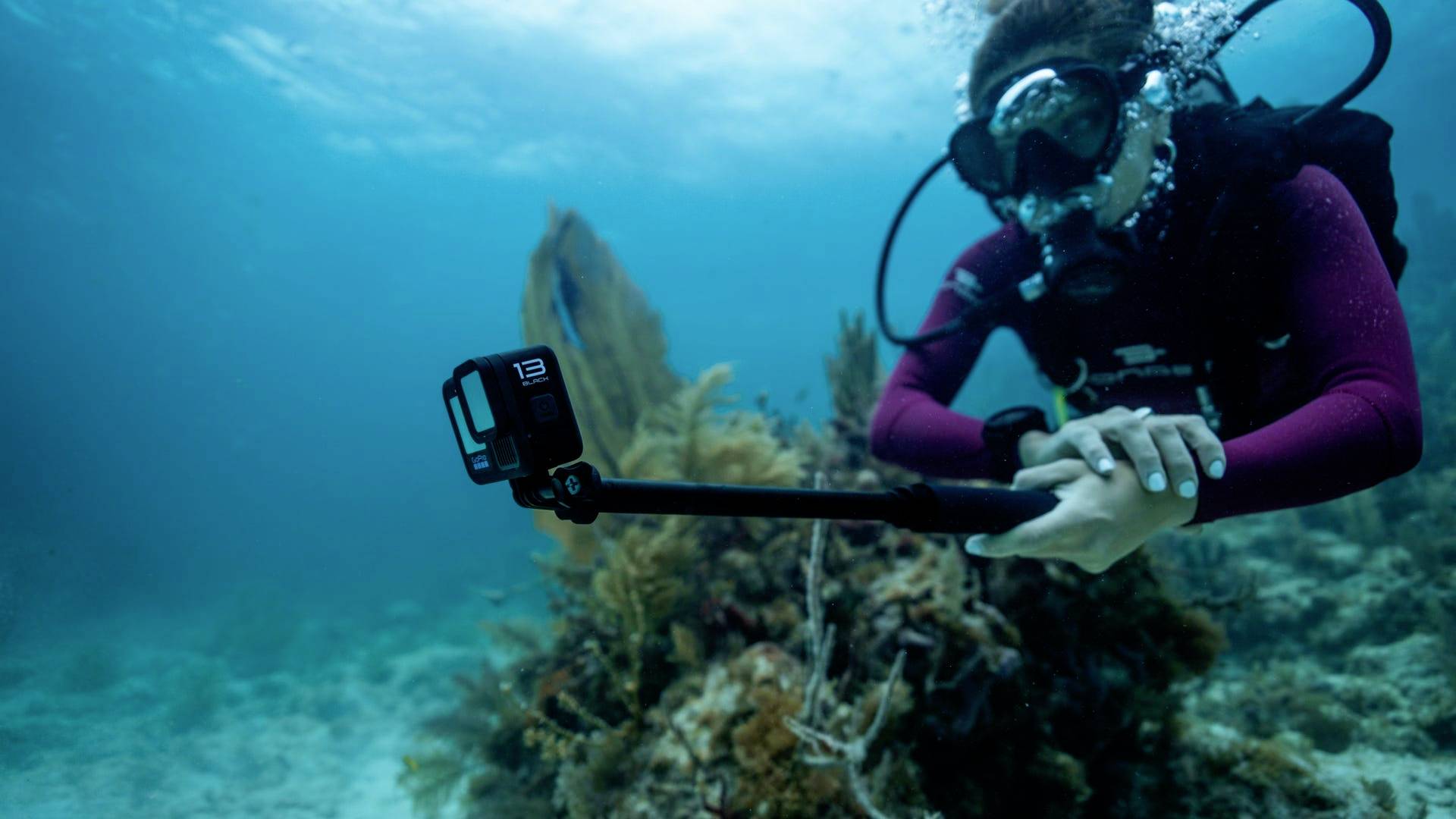 Een duikster onder water filmt een koraalrif met een actioncamera aan een selfie-stick. Ze draagt een duikbril en ademapparatuur.