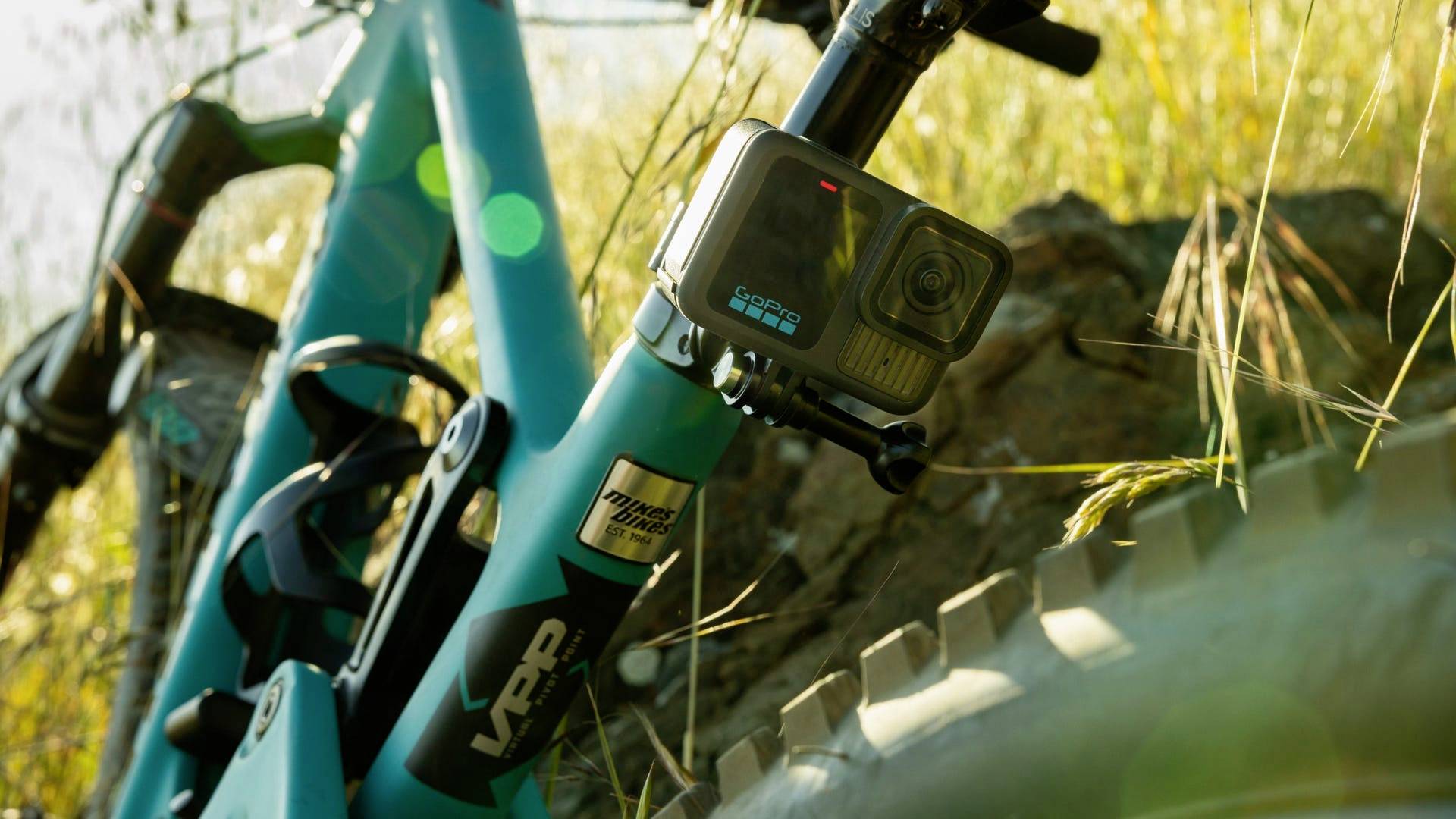 Een blauwe mountainbike met gemonteerde actiecamera staat op rotsachtig terrein, omringd door gras in het zonlicht.