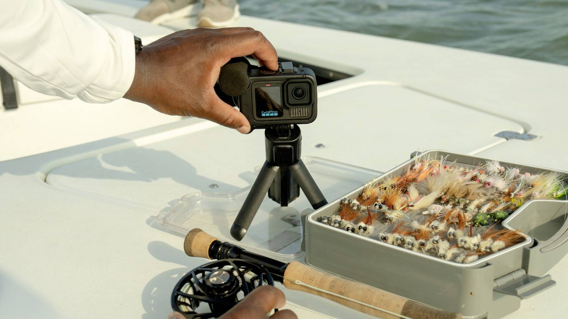 Een persoon bereidt een camera voor op een bootstoel. Naast de camera ligt een geopende doos met kunstvliegen voor het vissen.
