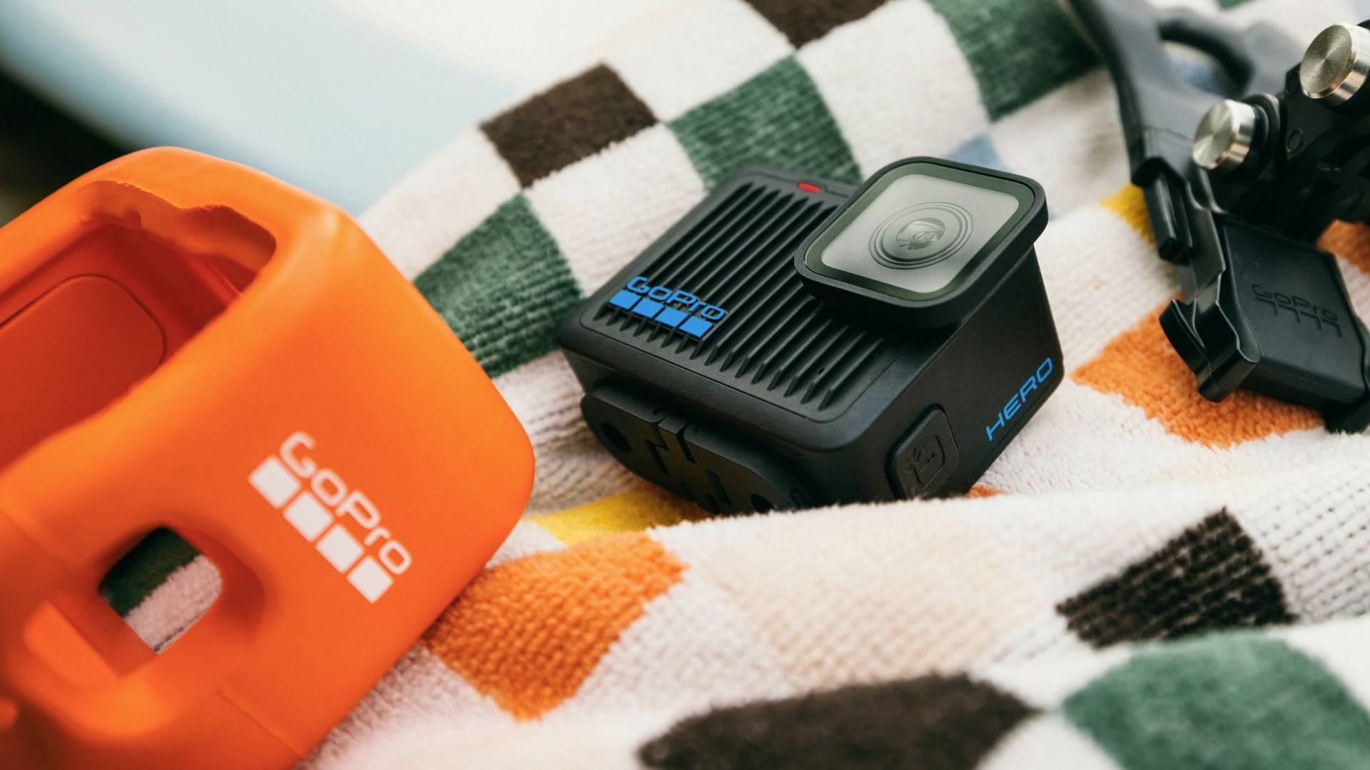 Compactcamera op gestreepte handdoek, samen met oranje houder en bevestigingstoebehoren. Ideaal voor buitenavonturen.