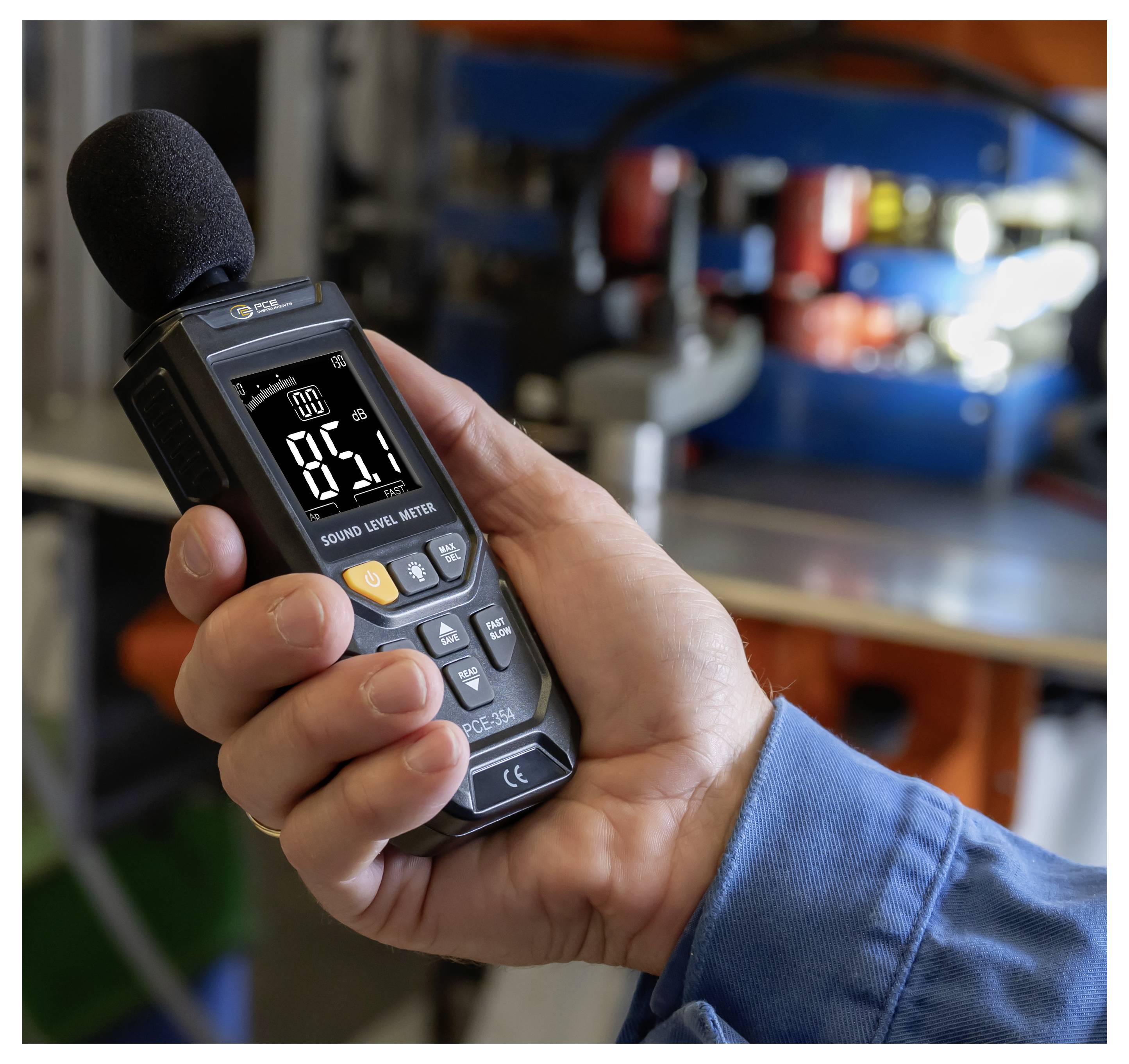 Hand die een geluidsniveaumeter met schuimrubberen microfoon vasthoudt, aflezend 85,1 dB, in een werkplaats- of industriële omgeving.